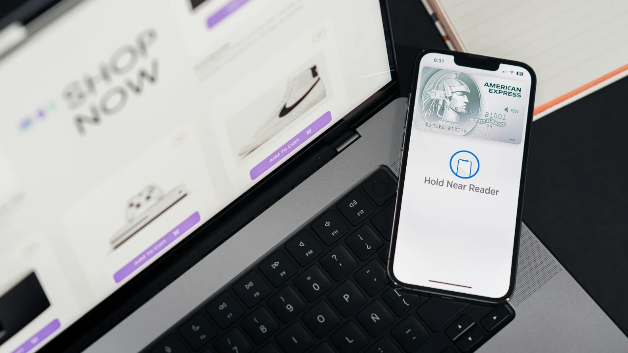 A laptop on a desk displaying an online checkout page next to a smartphone showing a contactless payment screen and a payment card.