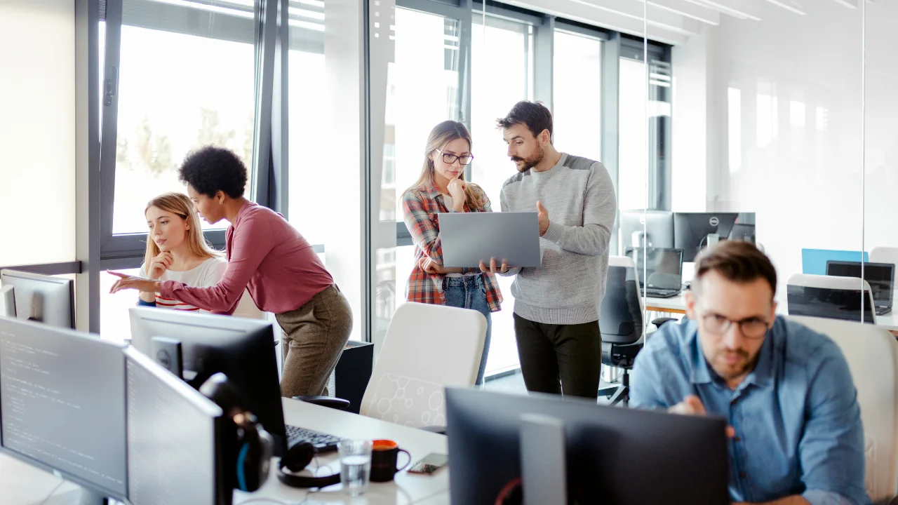 A diverse team of software developers and IT professionals working collaboratively on laptops and desktop computers in a bright, modern open-plan office.