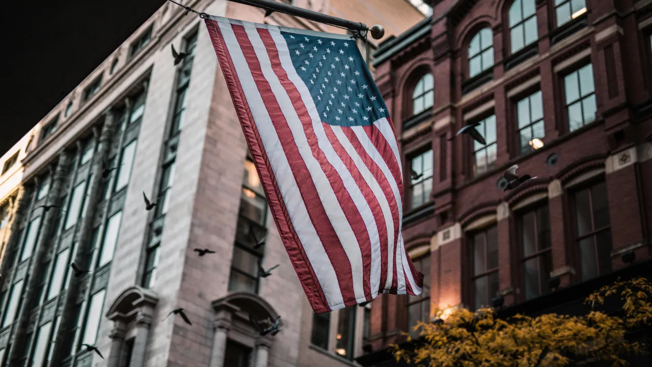 A low-angle shot of the American flag hanging from a flagpole in a historic U.S. city district with brick buildings and birds flying in the background.