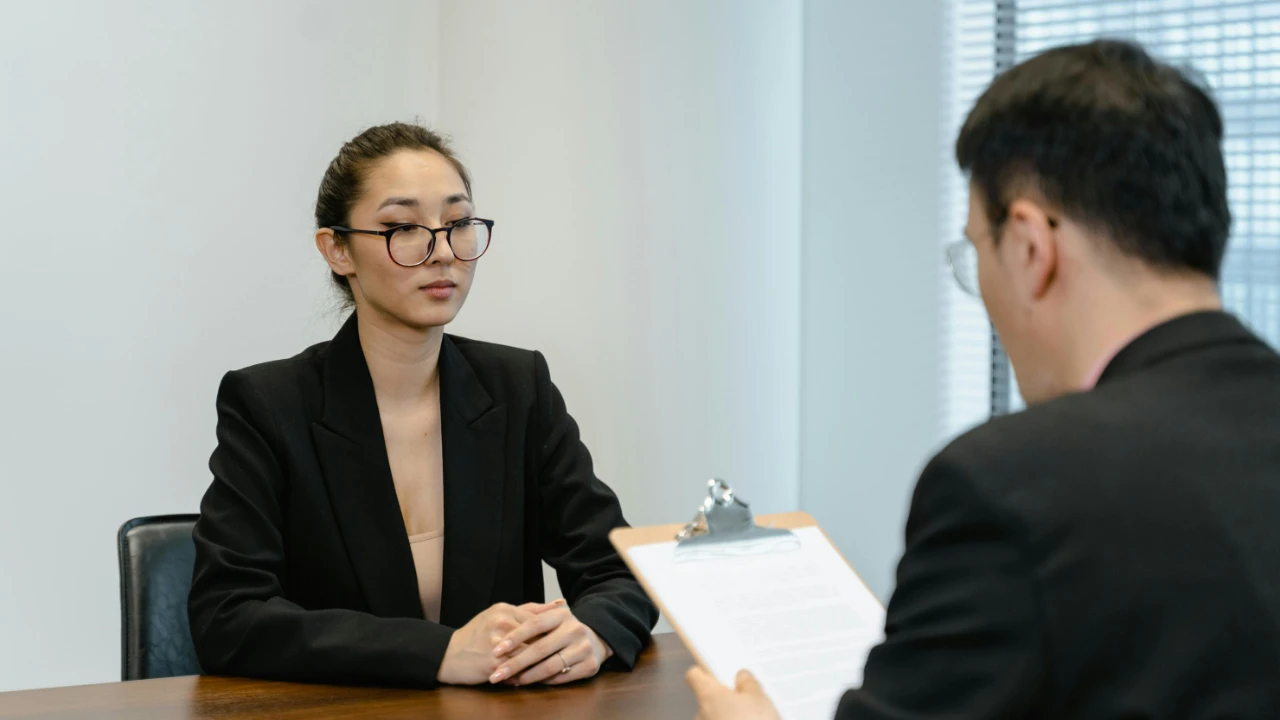 A professional woman in a black blazer sitting at a desk during a formal interview with a man holding a clipboard.