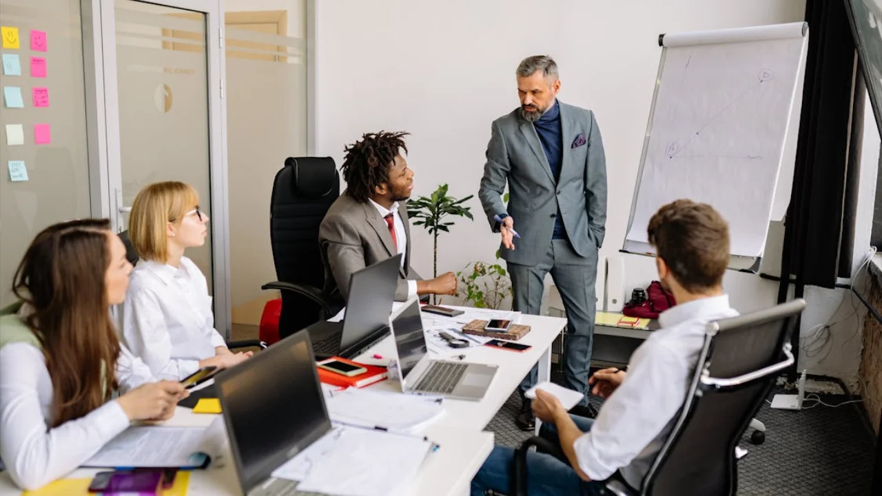 A senior executive in a formal suit stands at a whiteboard leading a strategy session with a diverse group of professional subordinates in a modern office.