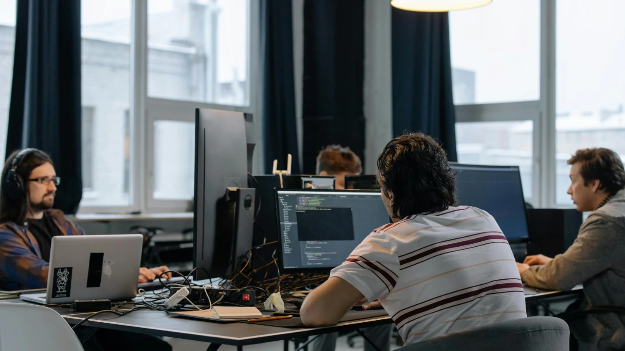 Several people seated at desks working on desktop computers in a modern office with large windows and overhead lighting.
