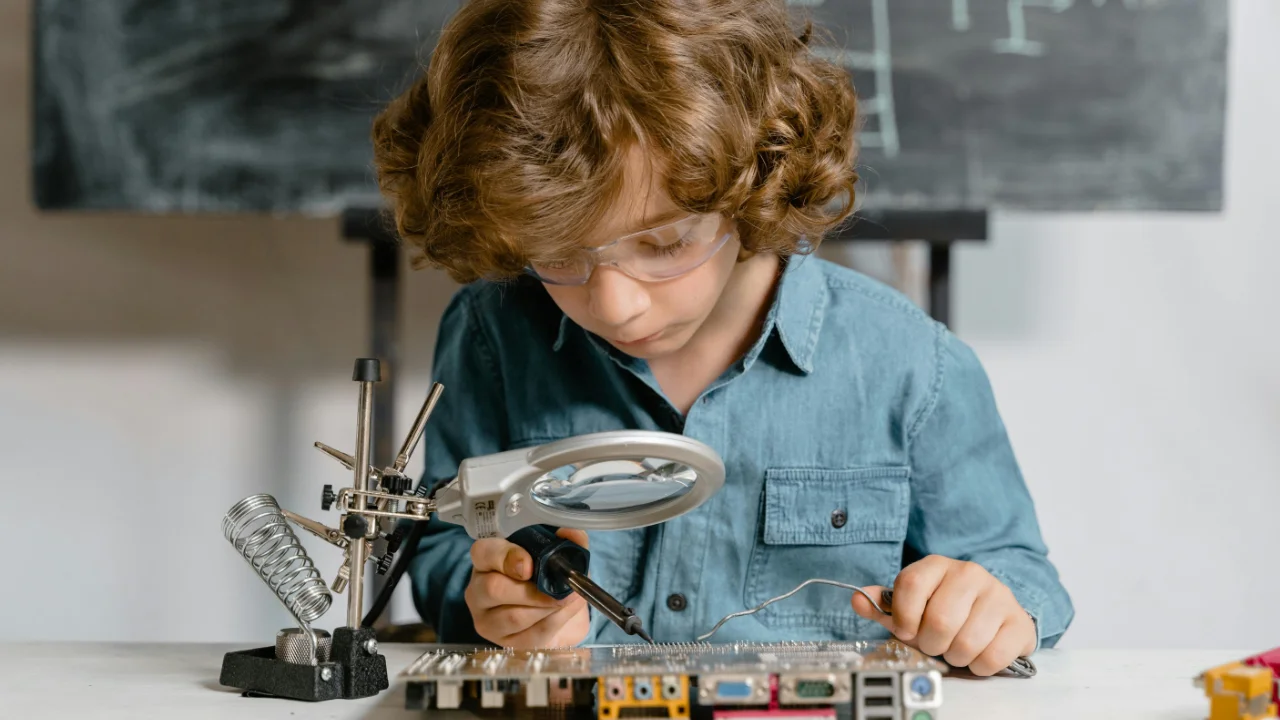 A young student wearing safety goggles and using a soldering iron to work on a complex computer motherboard, representing high-level technical skill and qualification.