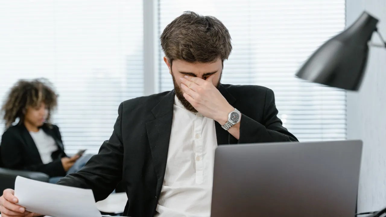 A stressed business professional sitting at a desk covering his face in frustration after learning his E-2 visa employment was terminated.