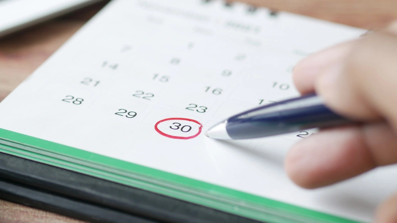 A close-up of a person's hand using a blue pen to circle the 30th on a calendar next to a calculator, representing L-1 visa timeline and extension planning.