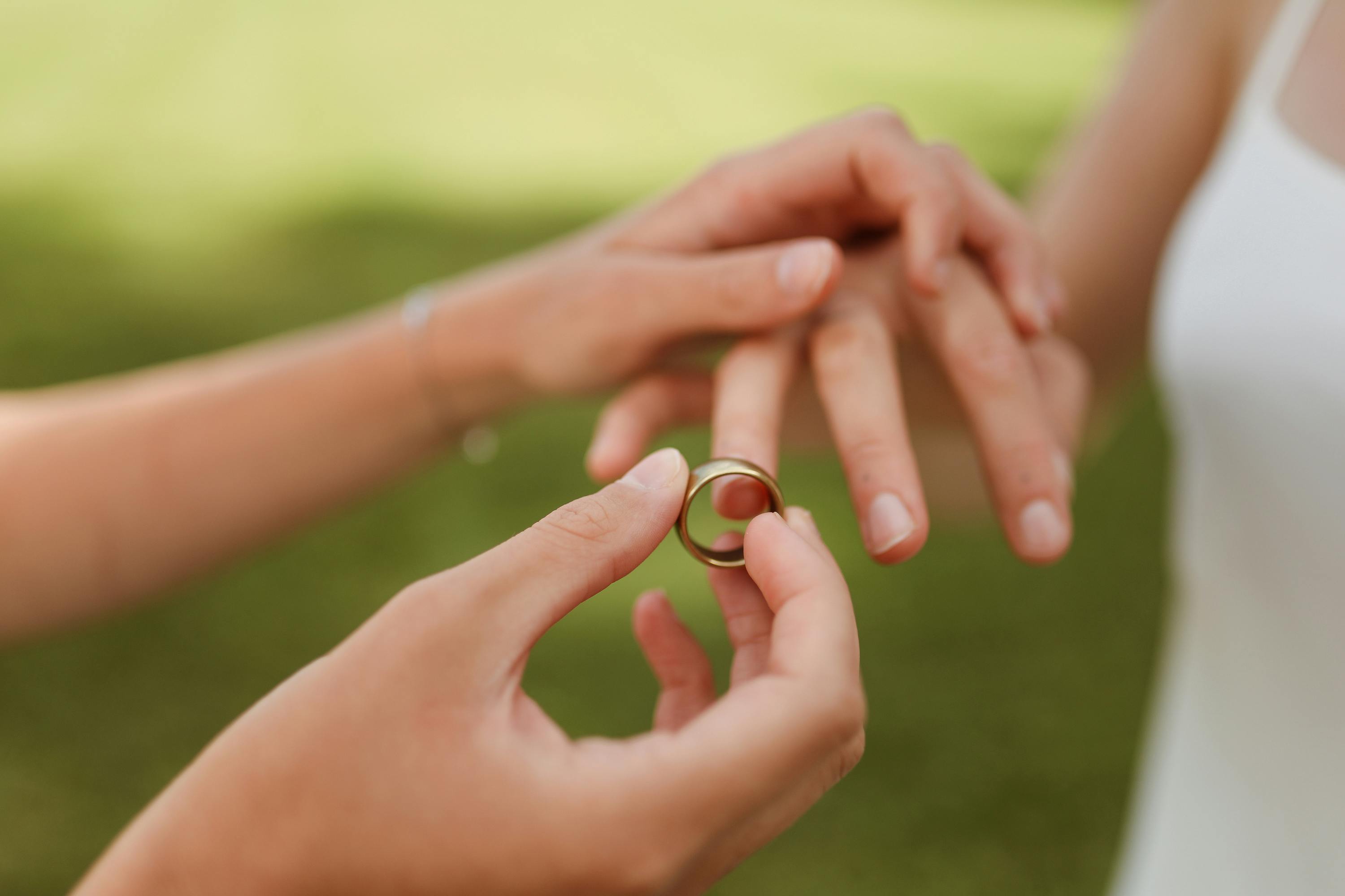Close-up of one hand placing a ring on another hand with a blurred green outdoor background