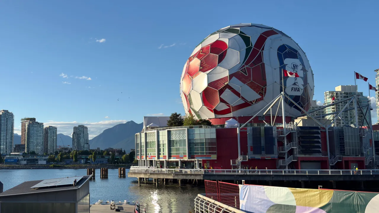 Large soccer-ball-shaped building flying a Canadian flag on the waterfront with mountains in the background, representing World Cup host cities.