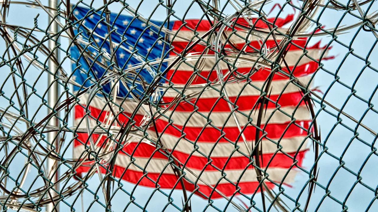 American flag seen through a chain-link fence topped with razor wire, illustrating admissibility vs. deportability in U.S. immigration law.