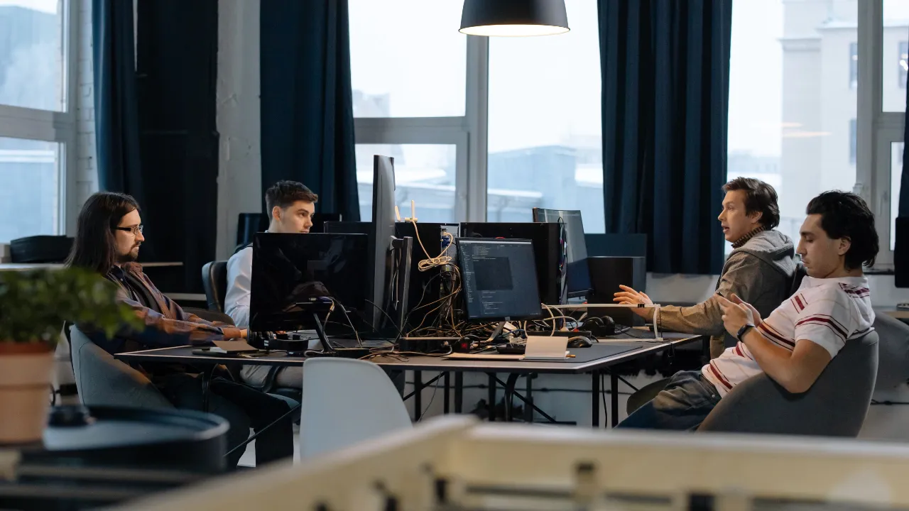 Several professionals working at computer workstations in a modern software development office with multiple monitors and collaborative workspace seating.
