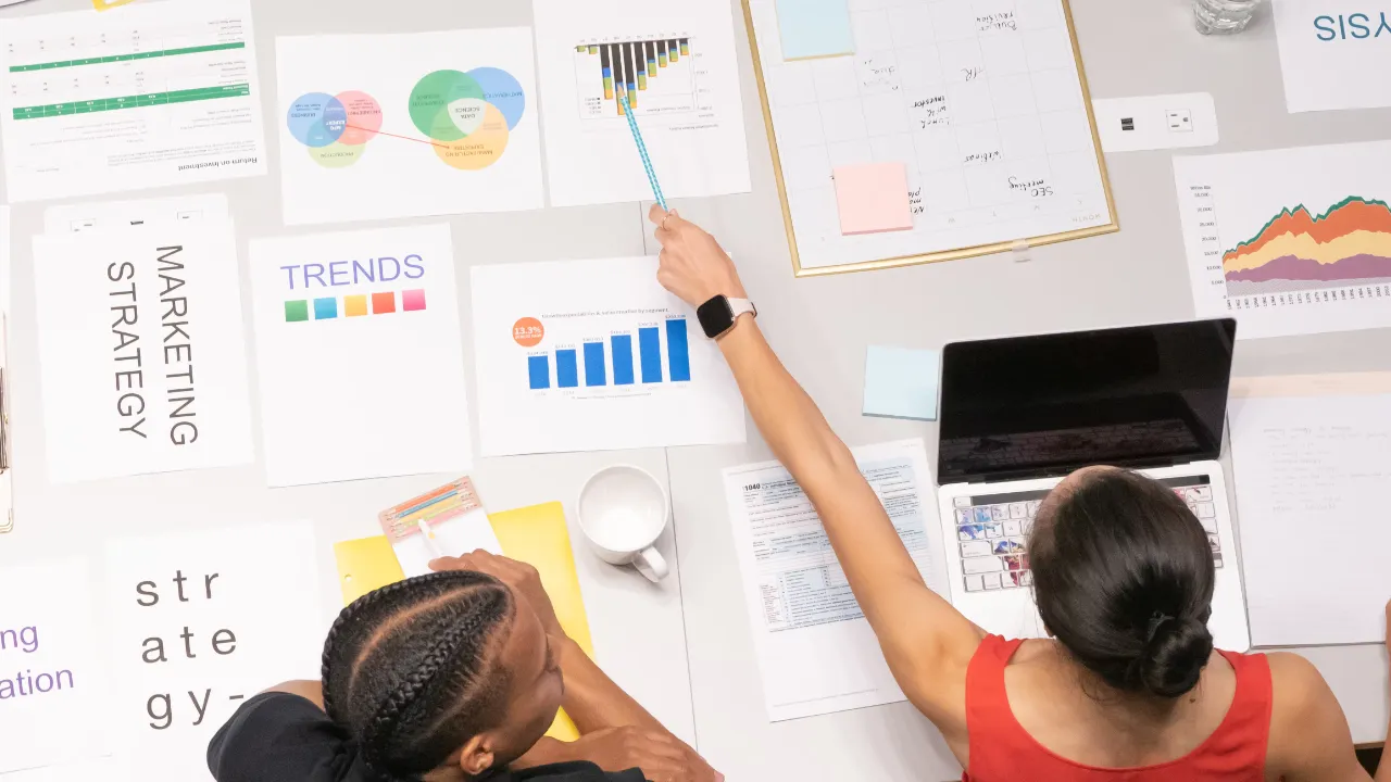 Overhead view of two professionals reviewing marketing strategy documents, charts, trend reports, and business planning materials on a conference table.