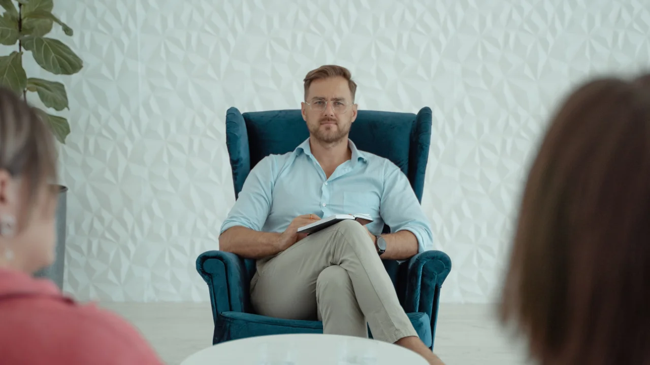 Professional seated in a blue armchair holding a notebook during a counseling session with two blurred participants visible in the foreground.