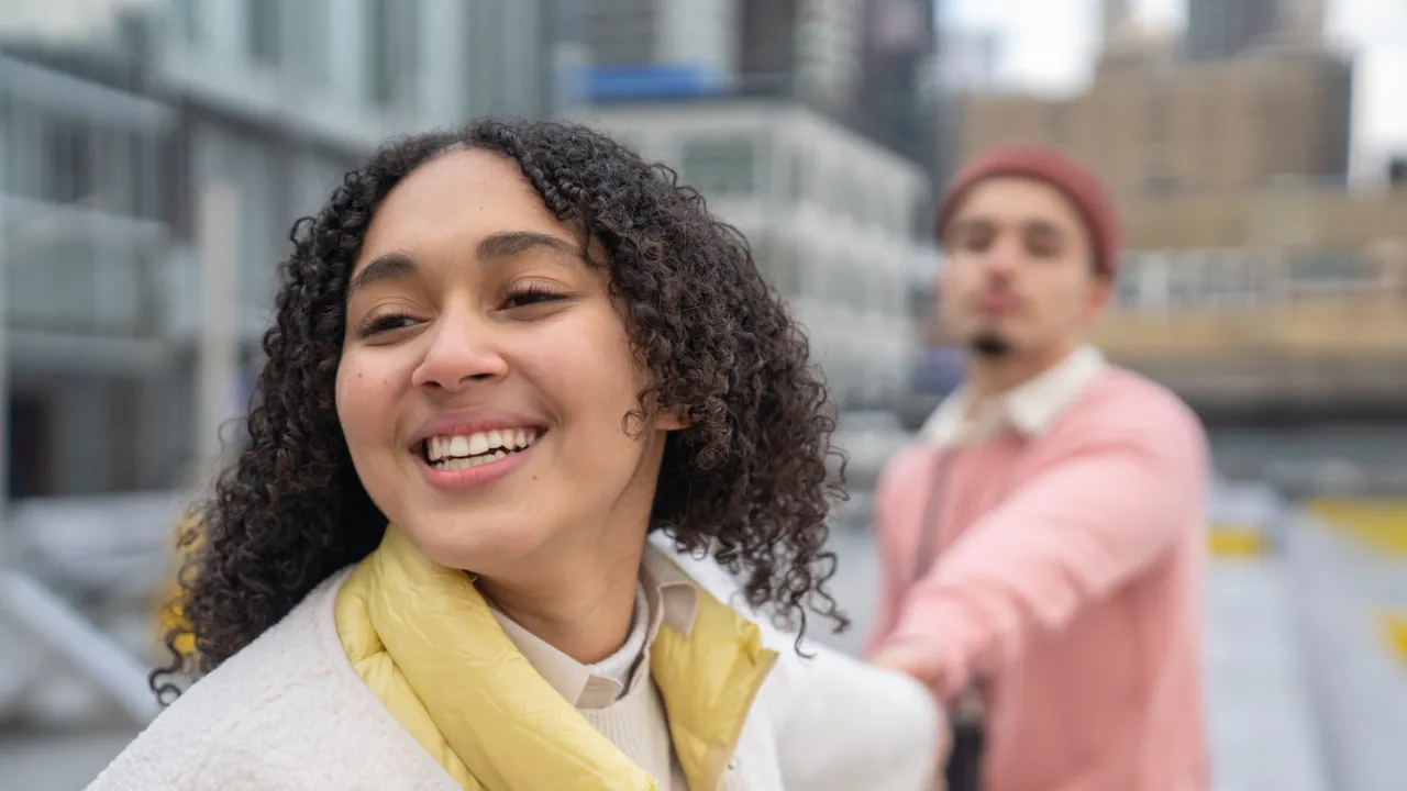 Smiling woman holding her partner's hand on a city walkway, representing a couple applying for adjustment of status as the spouse of a green card holder.