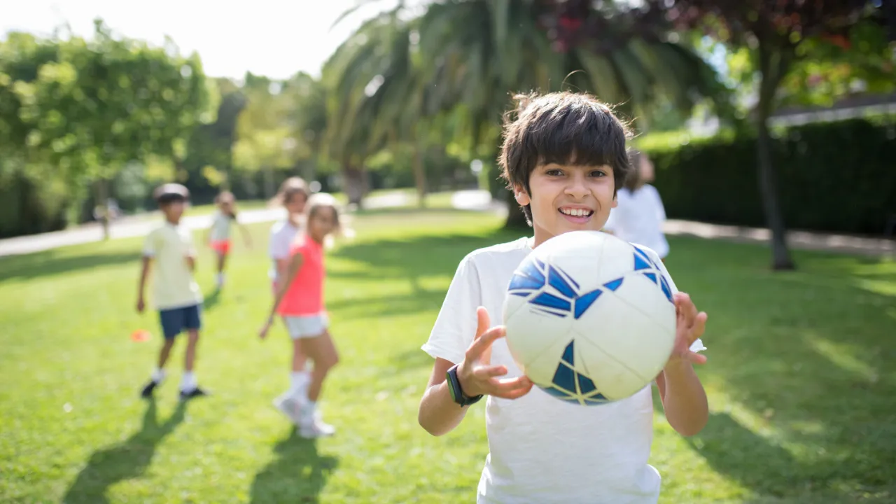 A smiling boy holds a ball on a grass field while other children play behind him