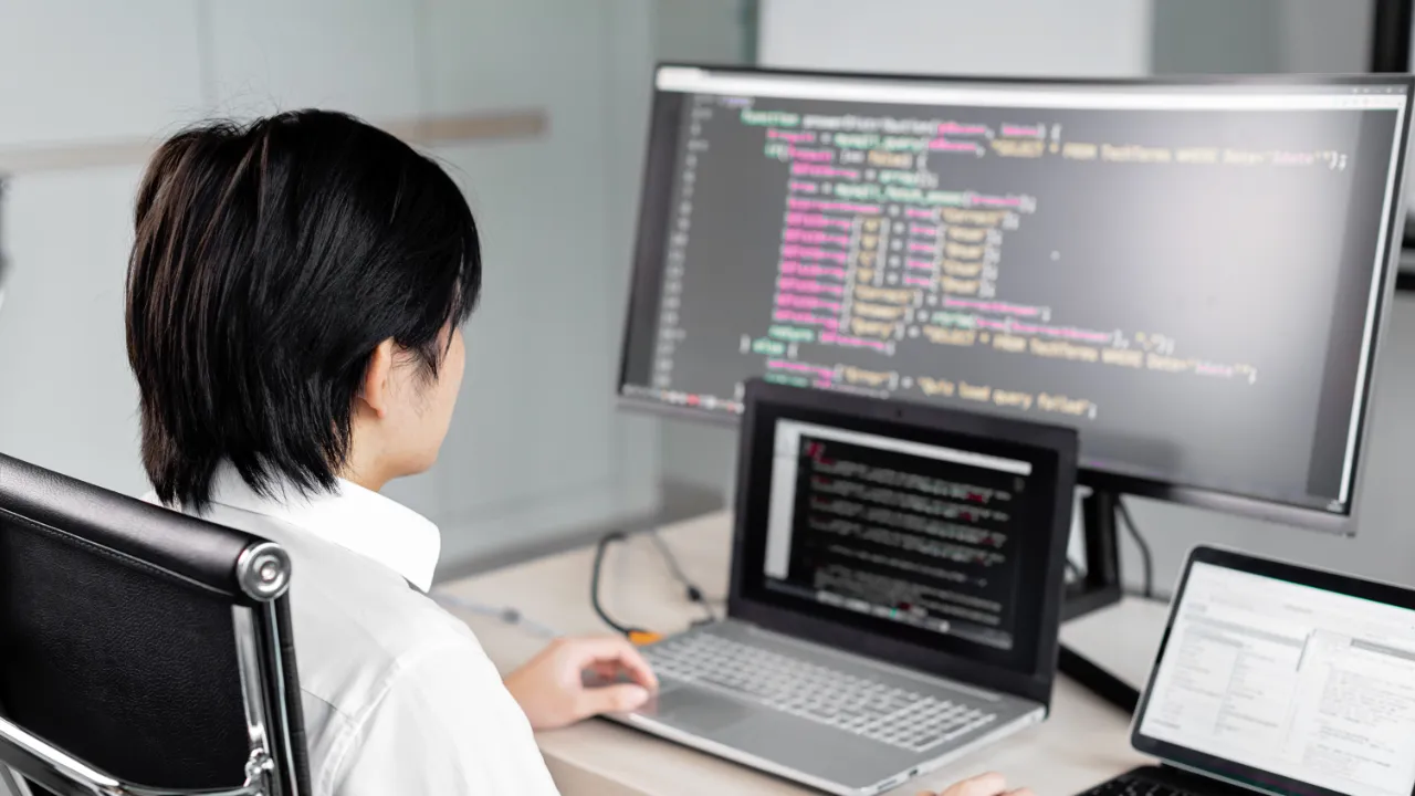 Person working at a desk with a laptop and monitor displaying software code in an office setting.