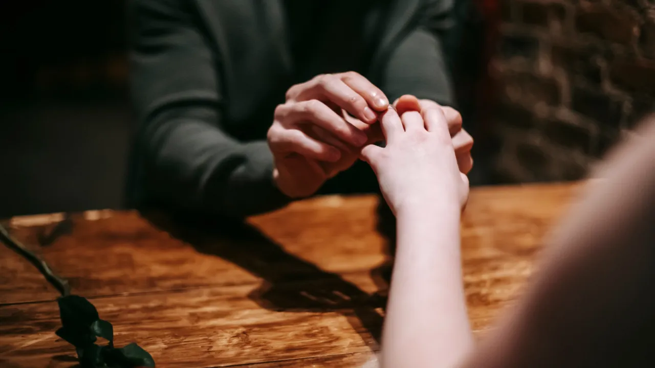 Close-up of a person placing an engagement ring on their partner's finger across a wooden table, with a red rose beside them