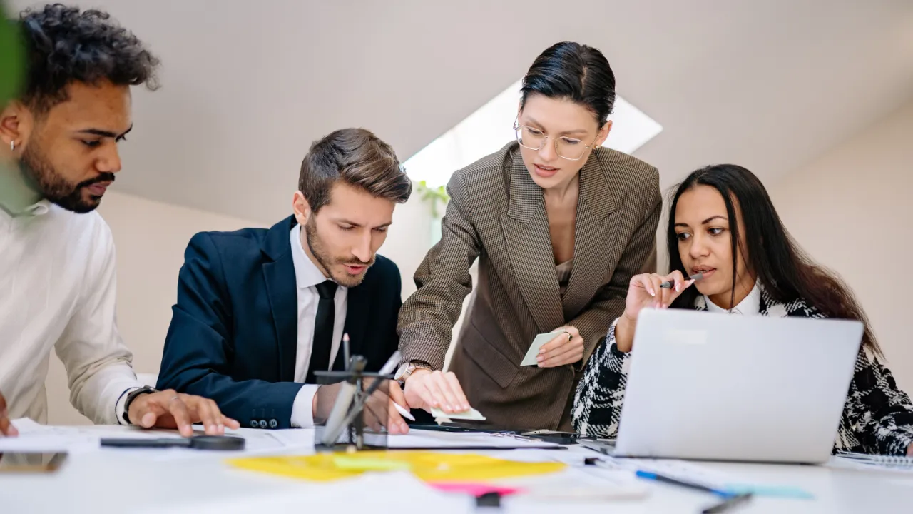 Four professionals gathered around a table reviewing documents and working on a laptop during a business consulting meeting.