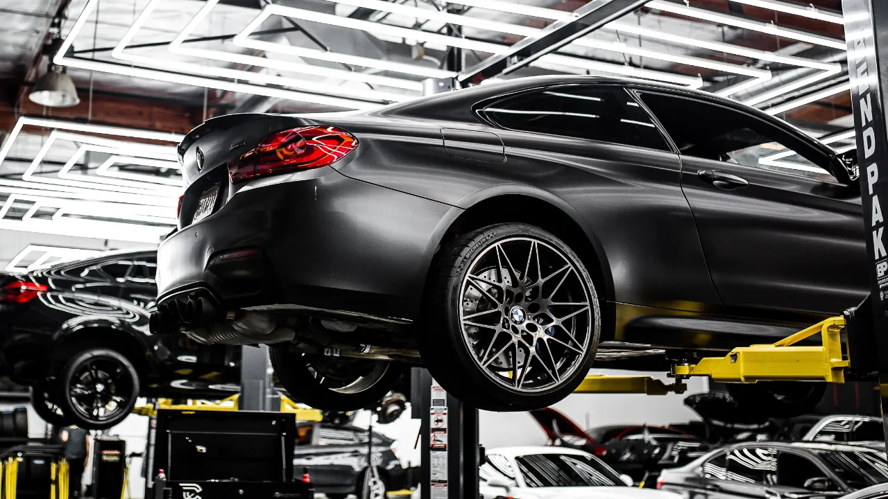 A car raised on a hydraulic lift inside a busy auto repair shop, with service bays and other vehicles in the background.