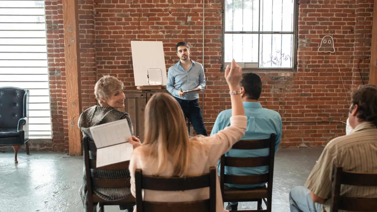 A coach leading a small group workshop in a brick-walled studio, with one participant raising a hand to ask a question.