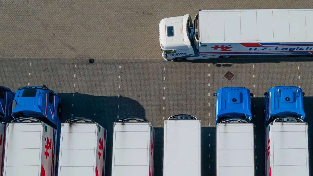 Overhead view of freight trucks parked in a row at a logistics depot, with one truck driving past the loading area.
