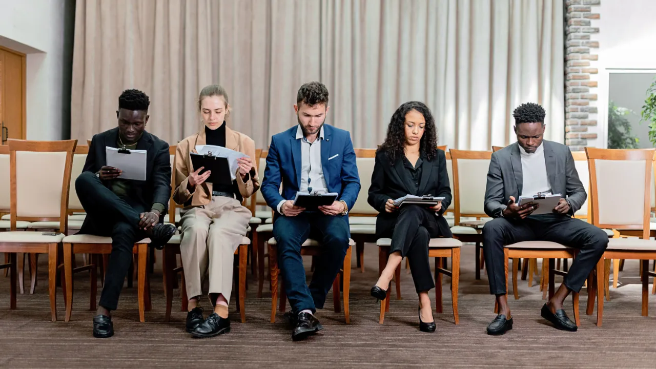 Five job candidates in business attire sit in a row of wooden chairs, each reviewing papers on a clipboard.