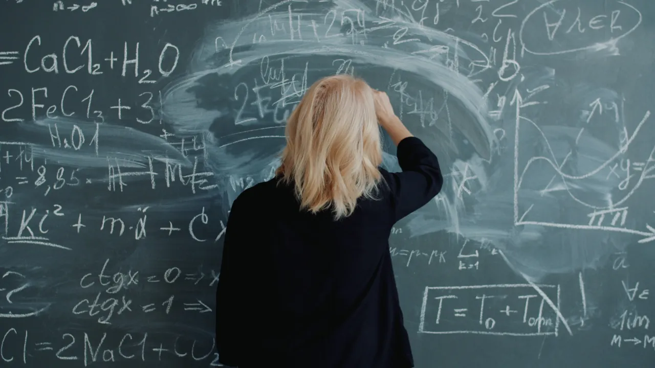 Person seen from behind facing a chalkboard covered in equations, one hand raised to their head