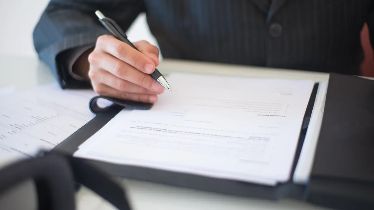 A person in a dark suit signing an immigration financial support form held in a black folder
