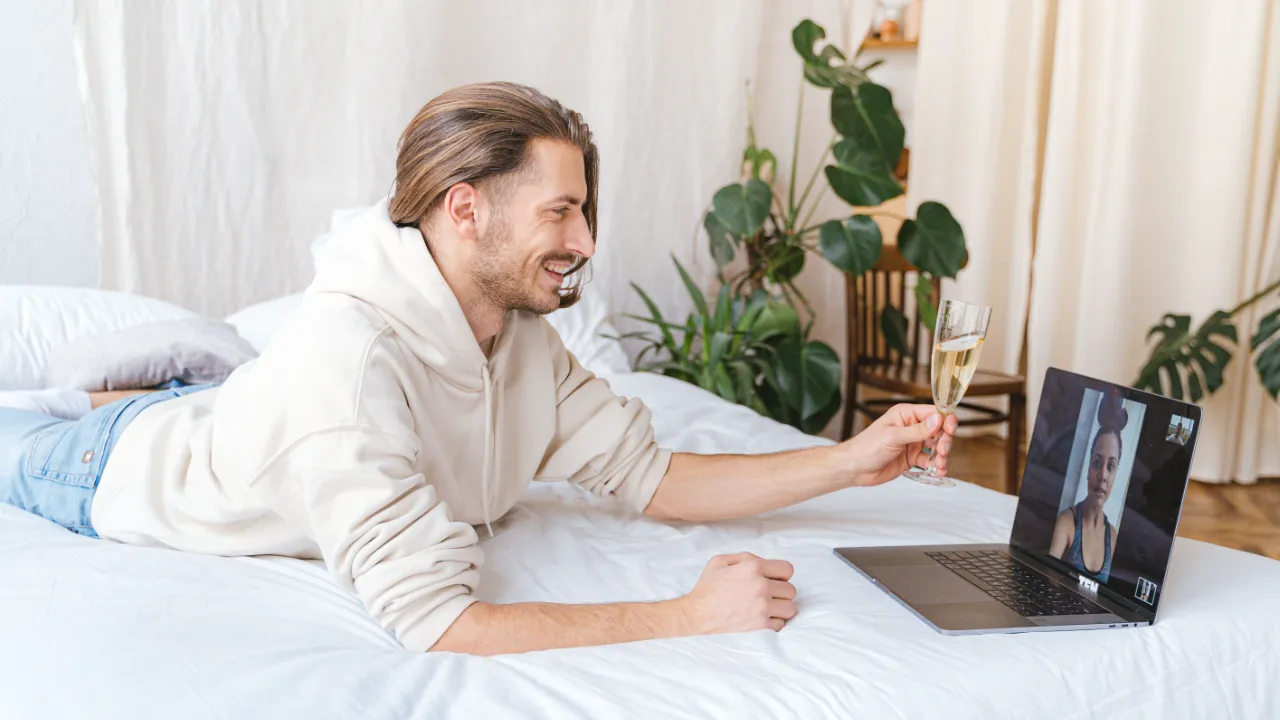 A man lying on a bed raising a glass during a video call with his partner on a laptop screen.