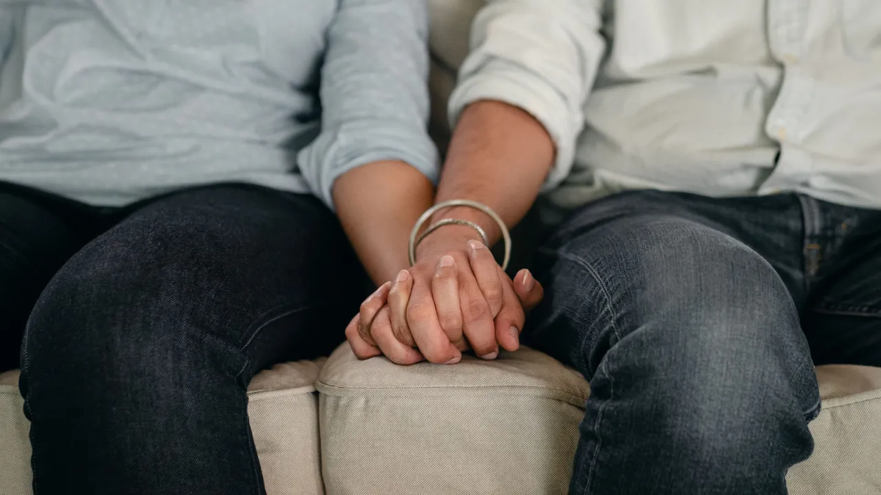 Two people sitting side by side on a beige sofa, holding hands, with their faces out of frame