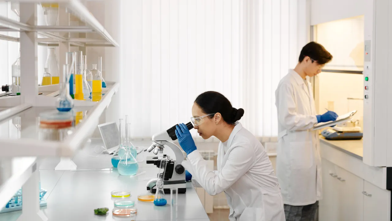 Two researchers working in a laboratory, one at a microscope and one taking notes at a fume hood.
