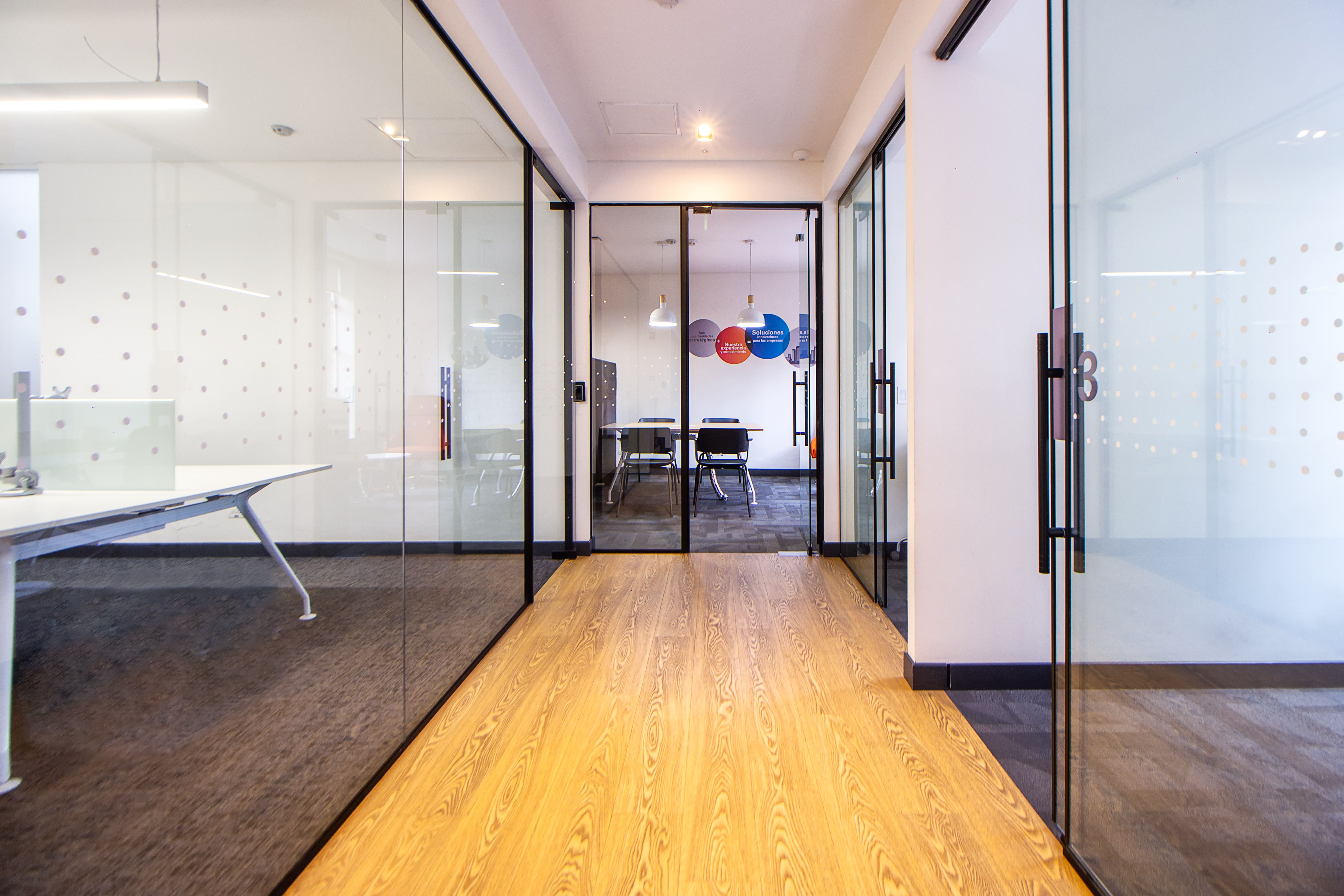 Office corridor lined with glass partition walls and black frames, leading to a meeting room with a table and chairs