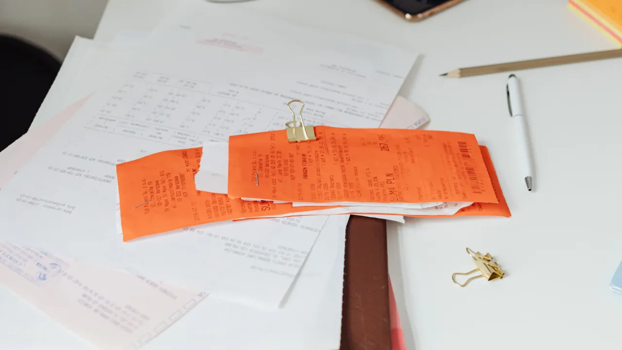 Desk covered with receipts, printed forms and paperwork clipped together beside a pen and notepad