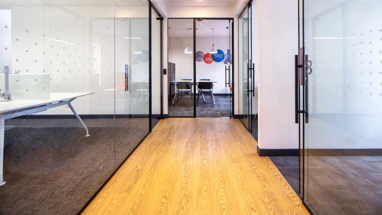 Office corridor lined with glass partition walls and black frames, leading to a meeting room with a table and chairs