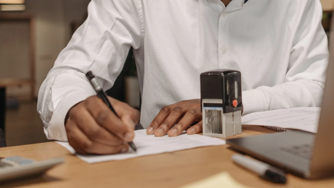Man in a white shirt filling out paperwork at a desk beside a self-inking stamp and an open laptop