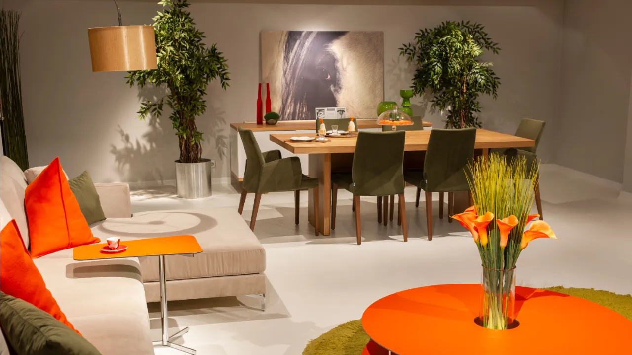 Staged open-plan living and dining room with a beige sectional sofa, orange cushions, a round orange coffee table and a wooden dining table with green upholstered chairs.