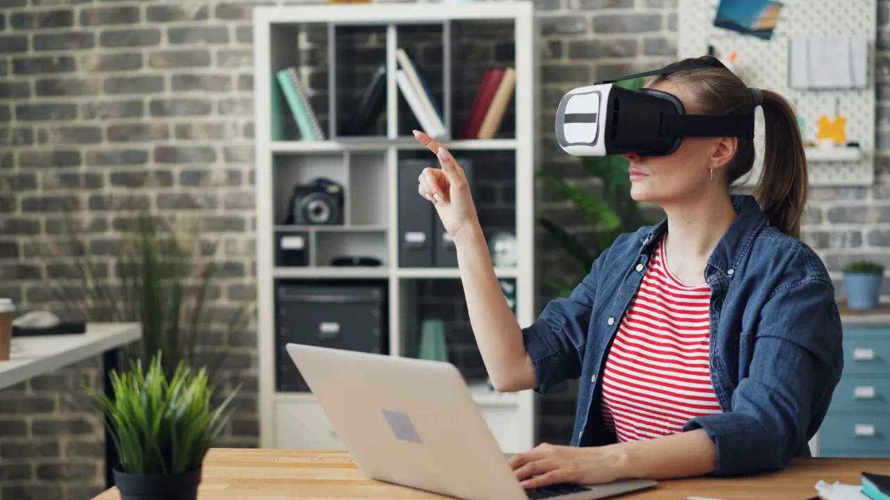 Woman wearing a virtual reality headset gestures in the air while typing on a laptop at a wooden desk in a brick-walled office.