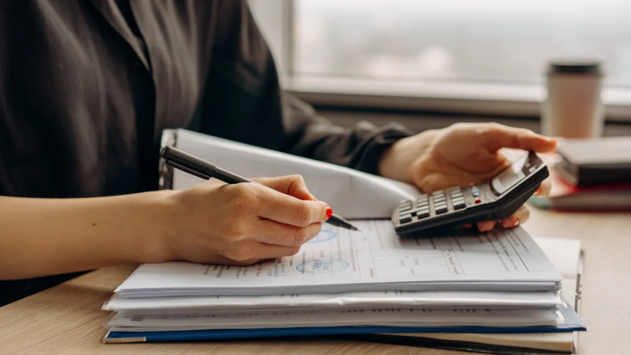 Hands holding a pen and a calculator over a stack of stamped financial documents and binders on a wooden desk by a window.