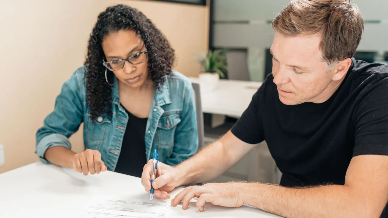 A man and a woman seated at a white table reviewing a printed form together, with the man pointing to a line with a pen.