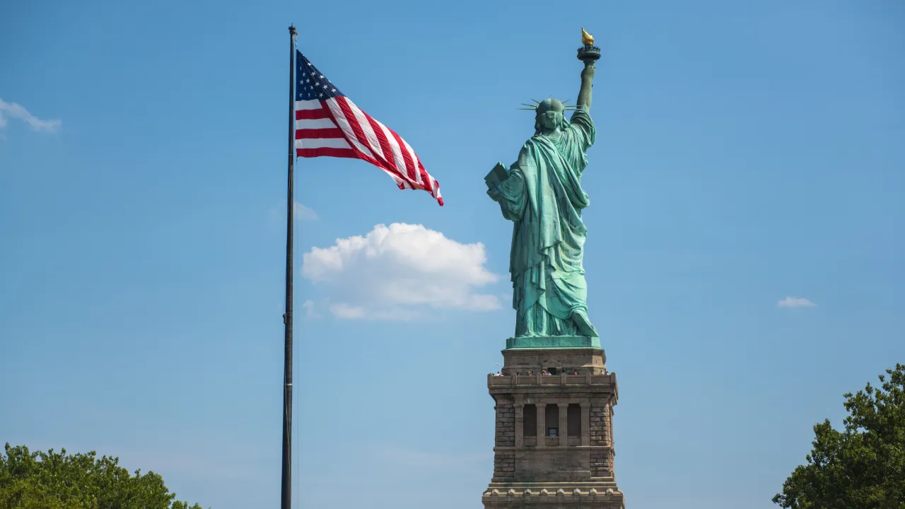 Statue of Liberty seen from behind next to an American flag waving under a clear blue sky