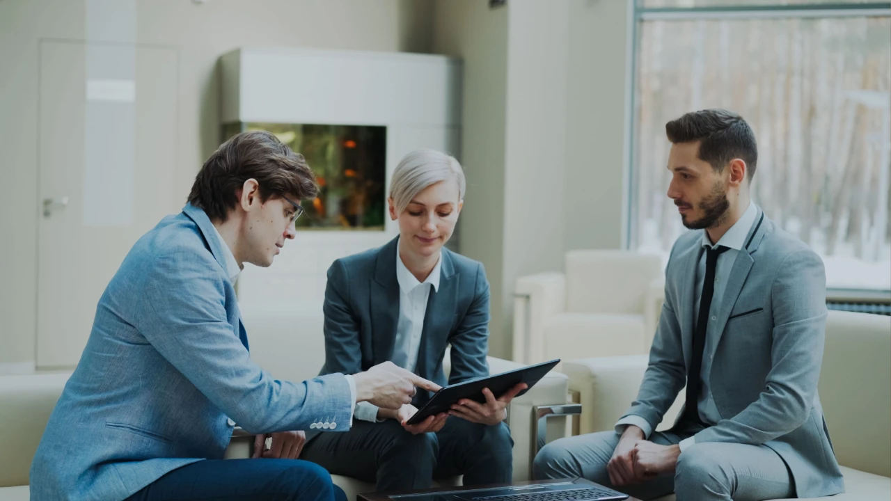 Three business professionals in a bright office lounge reviewing a tablet together during a consulting meeting