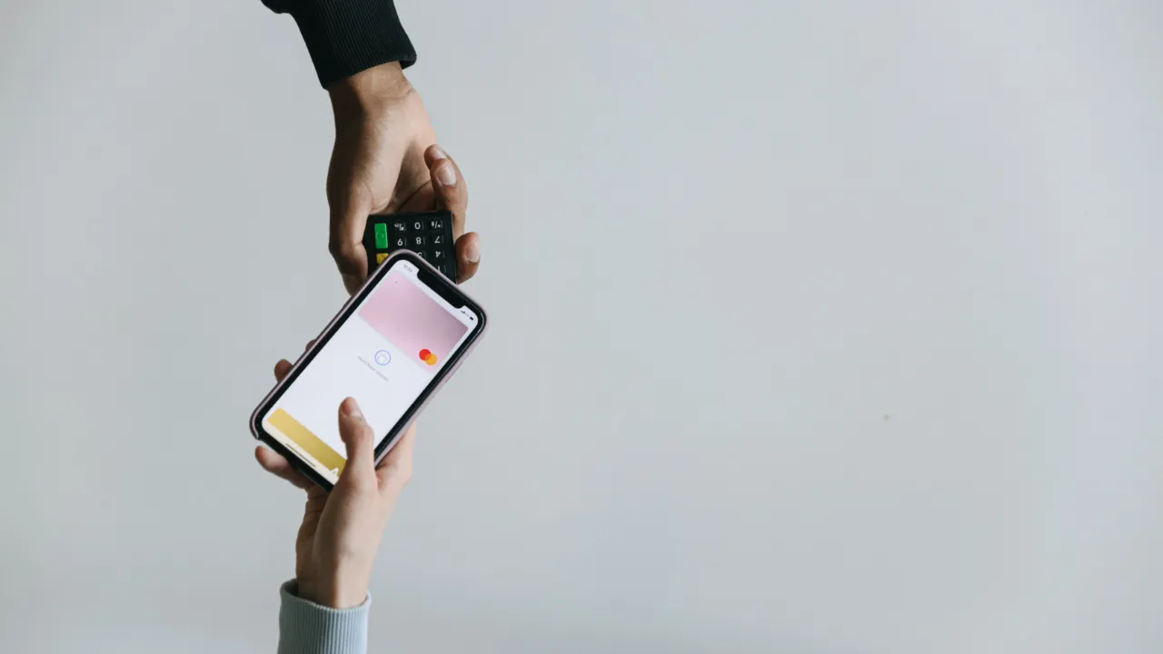 One hand holds a smartphone showing a digital wallet card near a card payment terminal held by another hand.